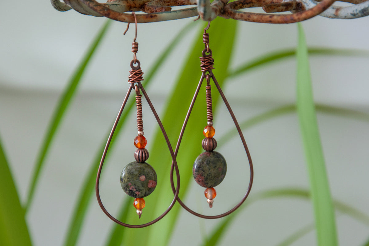Handmade copper teardrop drop earrings with unakite disc bead and carnelian accents, displayed on rustic metal stand with tropical greenery