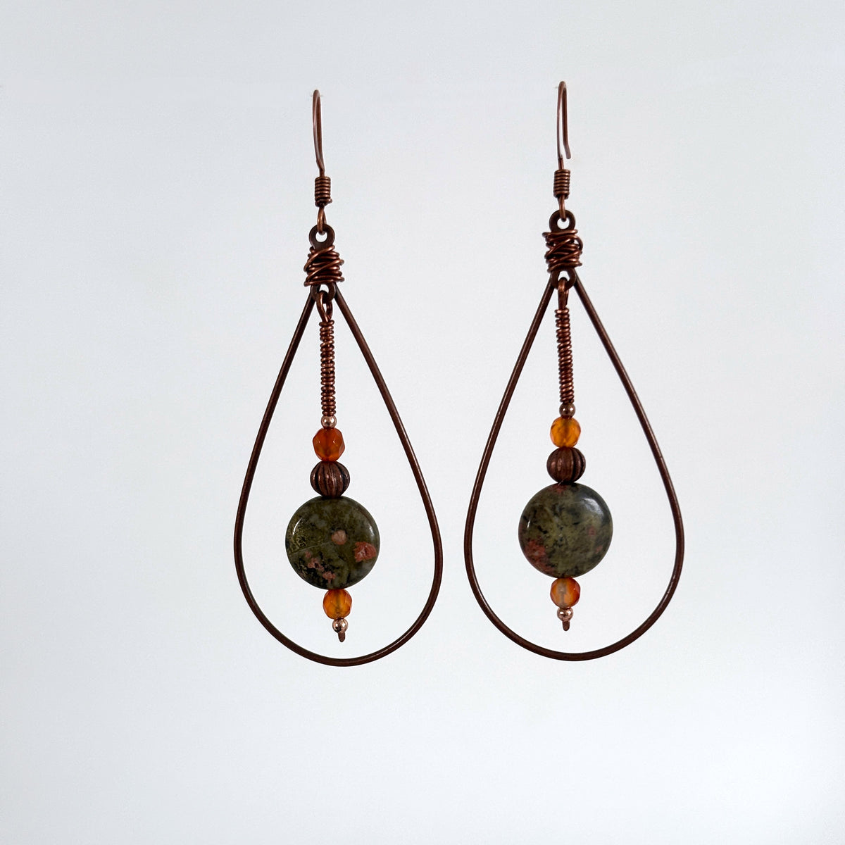 Handmade copper teardrop earrings with unakite disc bead, faceted carnelian, and wire-wrapped paddle drop, hanging against a neutral background