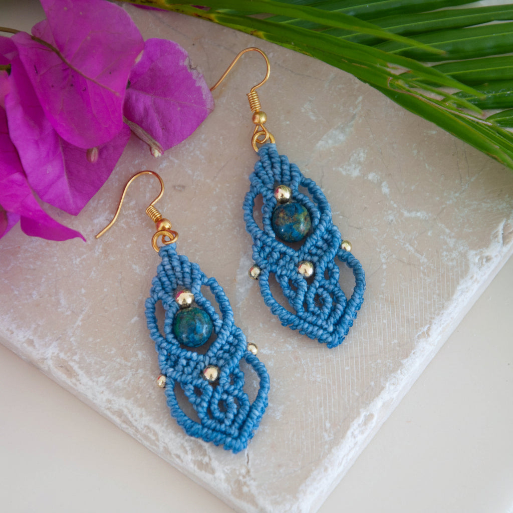 Pair of handmade slate blue macrame drop earrings with cascading lattice knotwork, 8mm round azurite gemstone center bead, 4mm and 3mm gold-tone metal beads, and gold-tone fishhook ear wires, laid on travertine tile with magenta bougainvillea and tropical palm leaf.