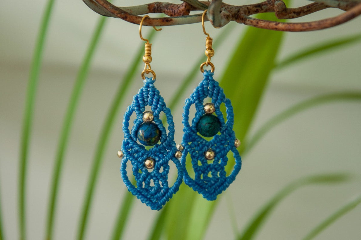 Pair of handmade slate blue macrame drop earrings with cascading lattice knotwork, 8mm round azurite gemstone center bead, 4mm and 3mm gold-tone metal beads, and gold-tone fishhook ear wires, hanging from rustic branch with blurred green foliage background.