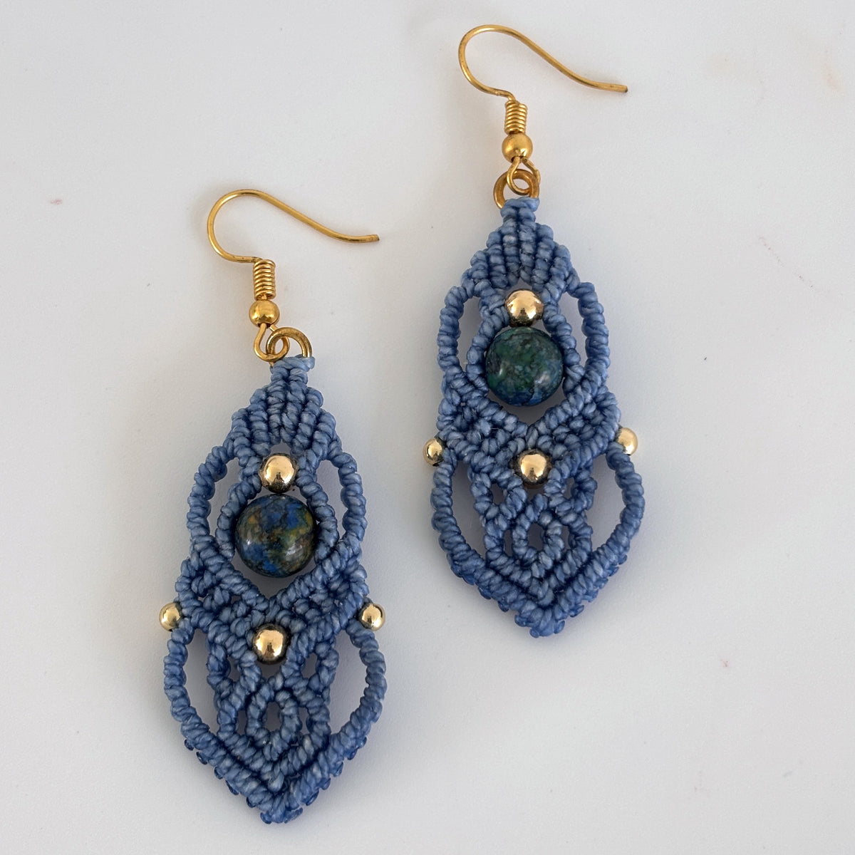 Pair of handmade slate blue macrame drop earrings with cascading lattice knotwork, 8mm round azurite gemstone center bead, 4mm and 3mm gold-tone metal beads, and gold-tone fishhook ear wires, displayed on white background showing full construction and bead detail.
