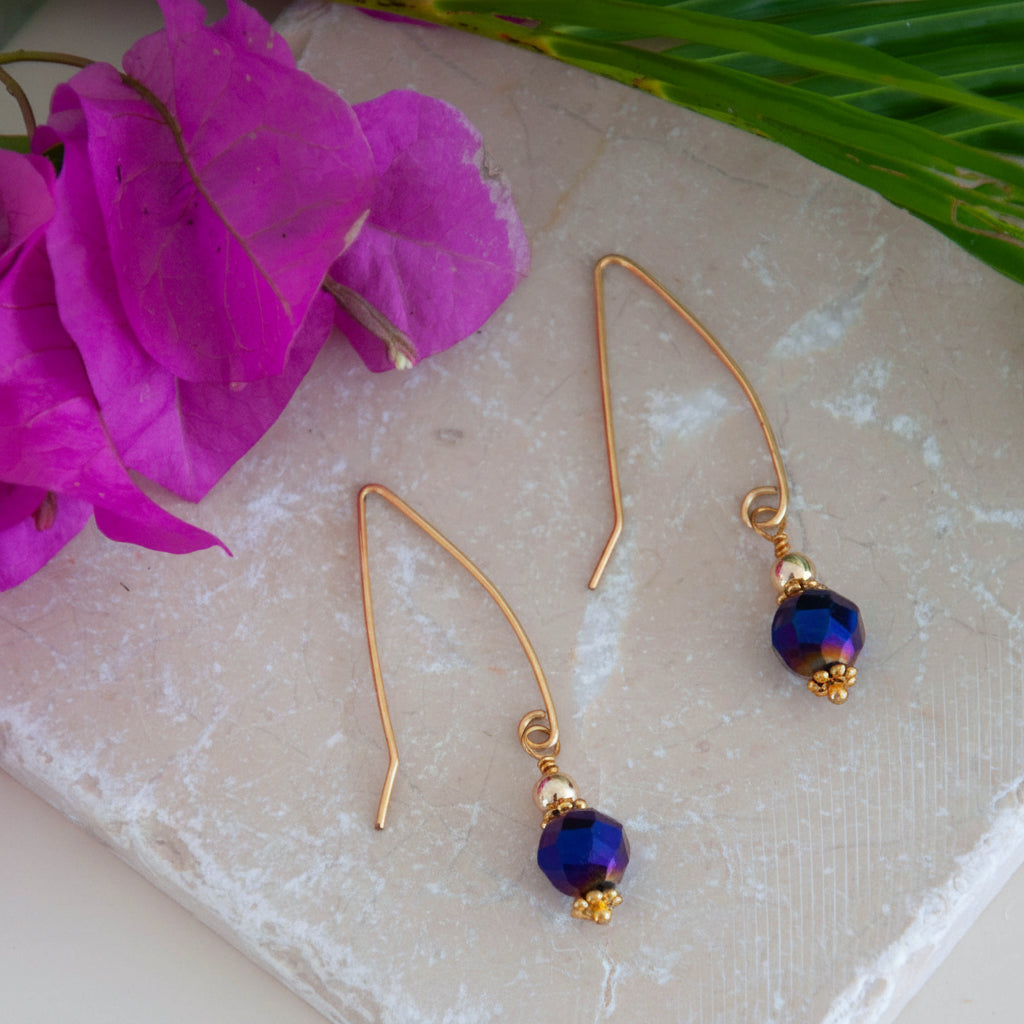 Cobalt blue vitrail glass drop earrings on travertine tile with magenta flowers and palm leaves.