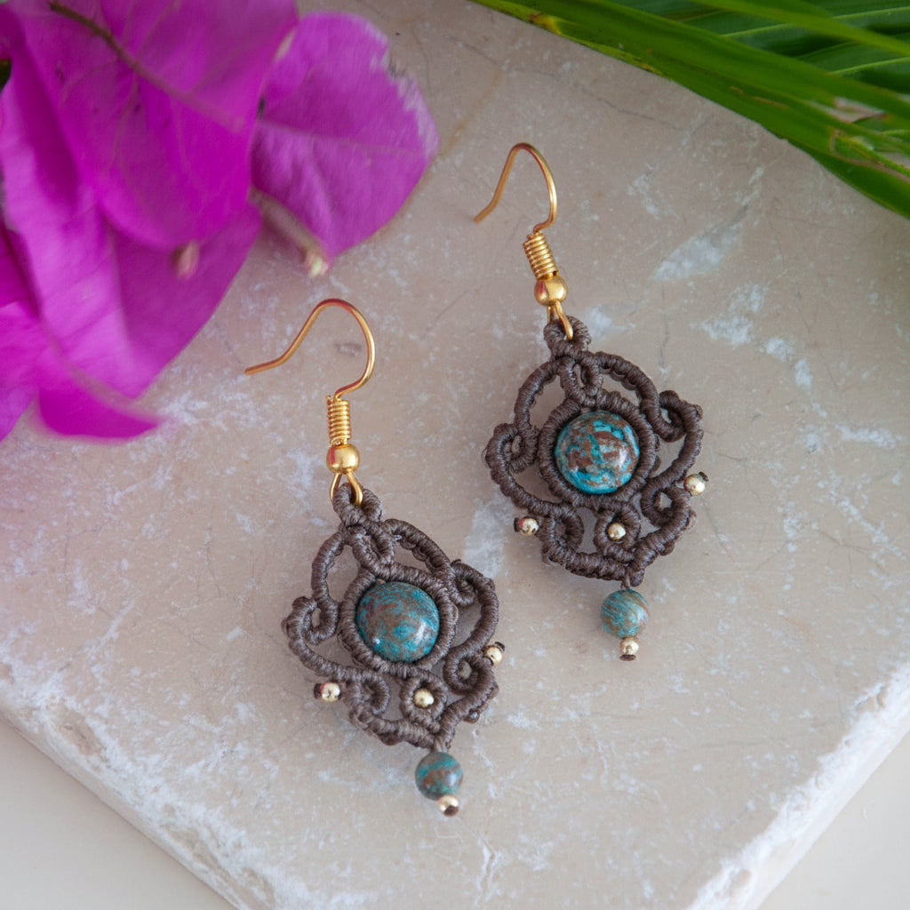 Handmade mocha brown macrame drop earrings with an 8mm blue calsilica bead, 2mm gold-tone accent beads, a 4mm blue calsilica drop bead, and gold-tone fishhook ear wires, laid on travertine tile with a magenta flower and tropical palm leaves.