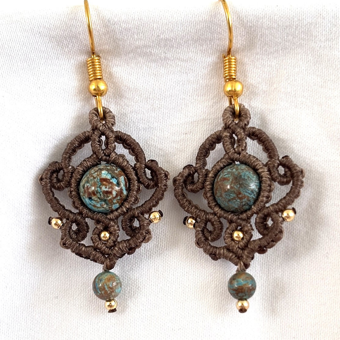 Pair of handmade mocha brown macrame drop earrings with ornate scrollwork silhouette, 8mm blue calsilica center beads, 2mm gold-tone accent beads, 4mm blue calsilica drop beads, and gold-tone fishhook ear wires, displayed on white fabric background.