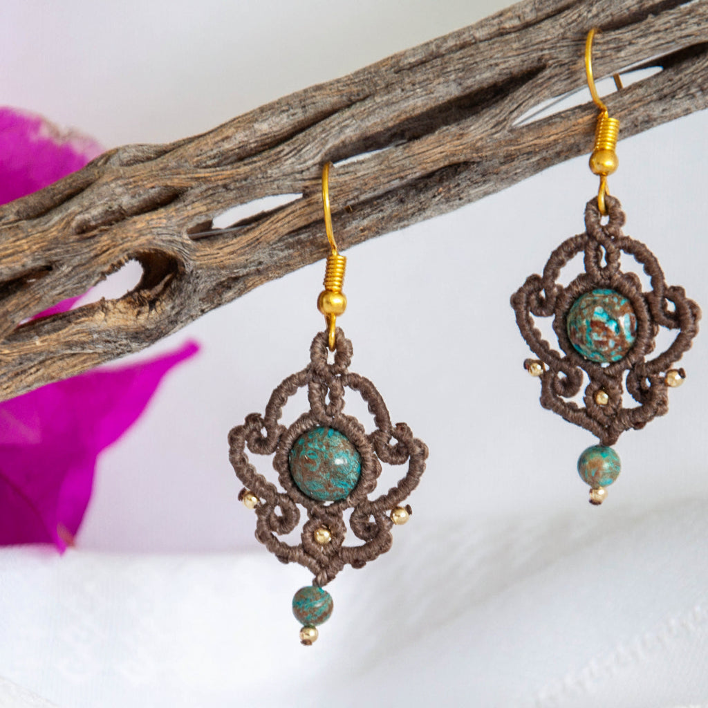 Handmade mocha brown macrame drop earrings with an 8mm blue calsilica bead, 2mm gold-tone accent beads, a 4mm blue calsilica drop bead, and gold-tone fishhook ear wires, hanging from driftwood with magenta bougainvillea.