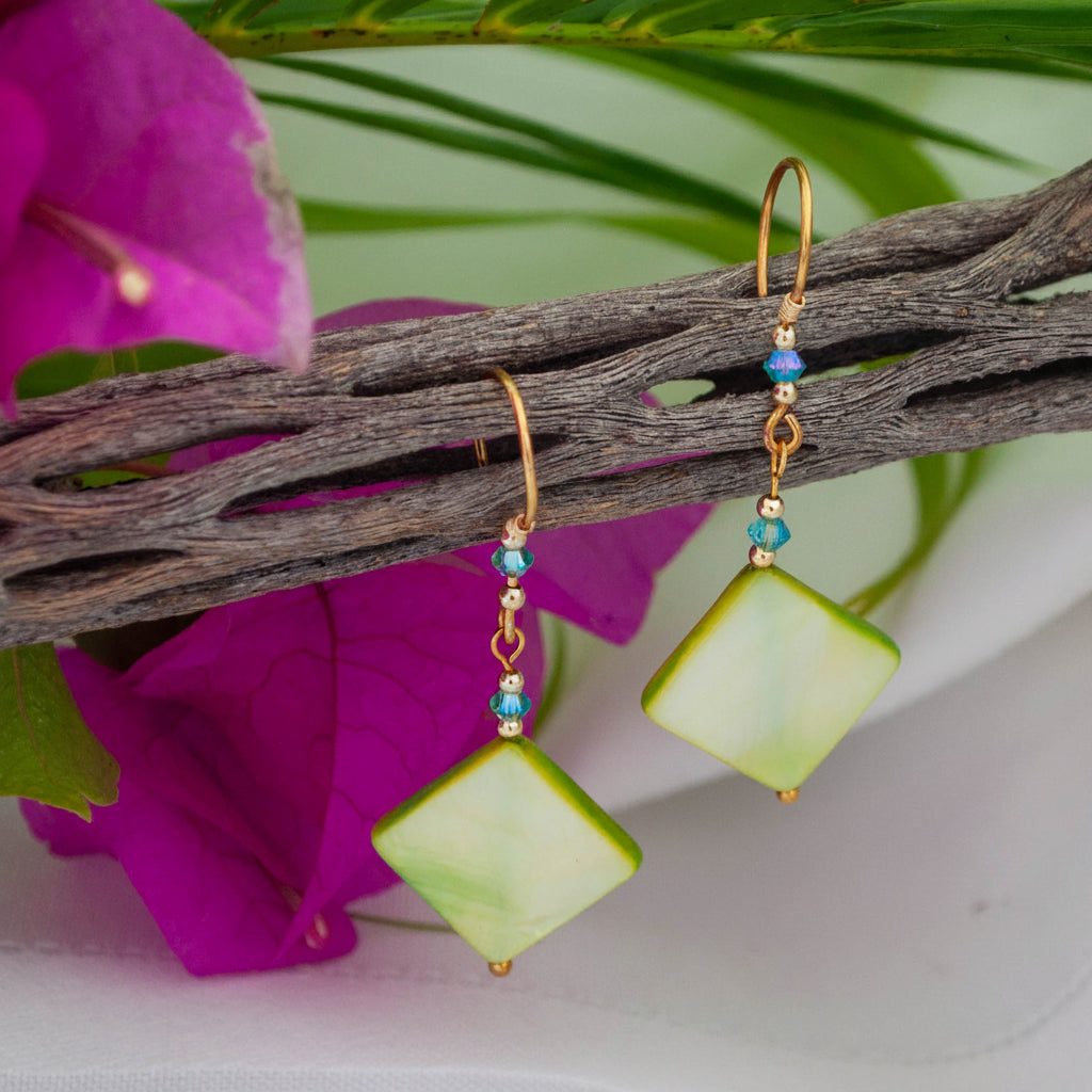 Handmade chartreuse mother-of-pearl diamond drop earrings with aqua Swarovski bicone crystals, gold-tone metal bead accents, brass wire wrapping, and handcrafted brass French wires, hanging from a driftwood branch with magenta bougainvillea and tropical palm leaves.