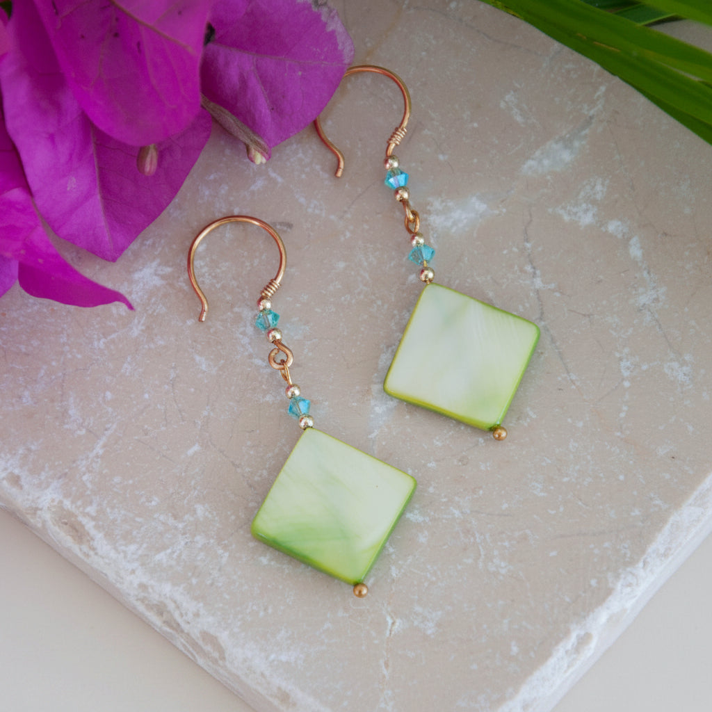 Handmade chartreuse mother-of-pearl diamond drop earrings with aqua Swarovski bicone crystals, gold-tone metal bead accents, brass wire wrapping, and handcrafted brass French wires, laid on a travertine tile with a magenta flower and tropical green leaves.