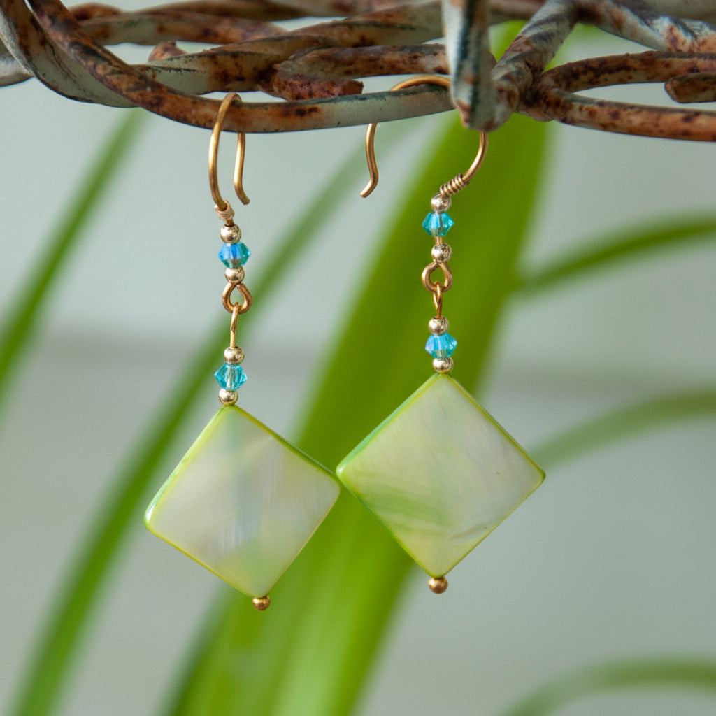 Handmade chartreuse mother-of-pearl diamond drop earrings with aqua Swarovski bicone crystals, gold-tone metal bead accents, brass wire wrapping, and handcrafted brass French wires, hanging from a rustic metal stand with tropical green leaves in the background.
