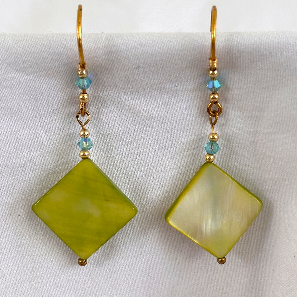 Pair of handmade chartreuse mother-of-pearl diamond drop earrings (15mm × 15mm) with 3mm aqua Swarovski bicone crystals, 2mm round gold-tone bead accents, brass wire wrapping, and handcrafted brass French wires, displayed on white fabric background showing natural iridescence.