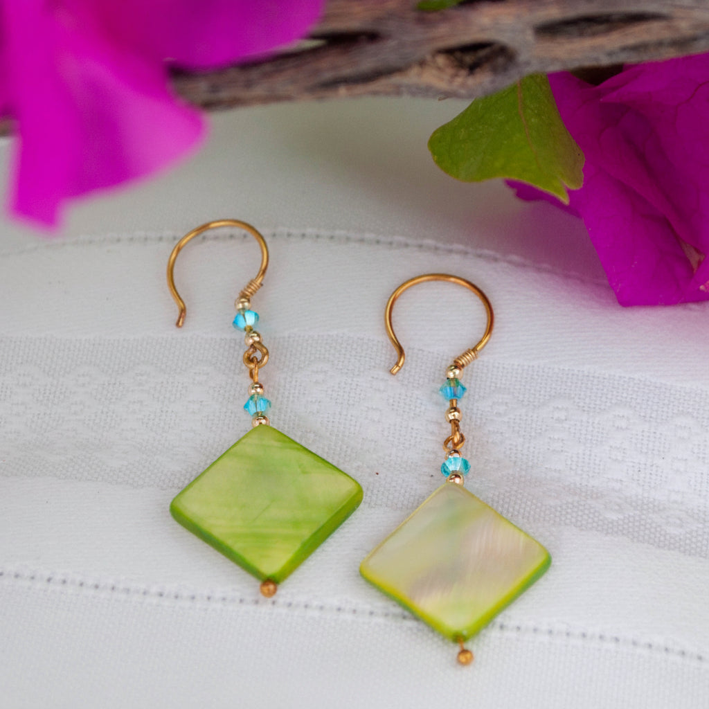 Handmade chartreuse mother-of-pearl diamond drop earrings with aqua Swarovski bicone crystals, gold-tone metal bead accents, brass wire wrapping, and handcrafted brass French wires, laid on white linen with magenta bougainvillea.