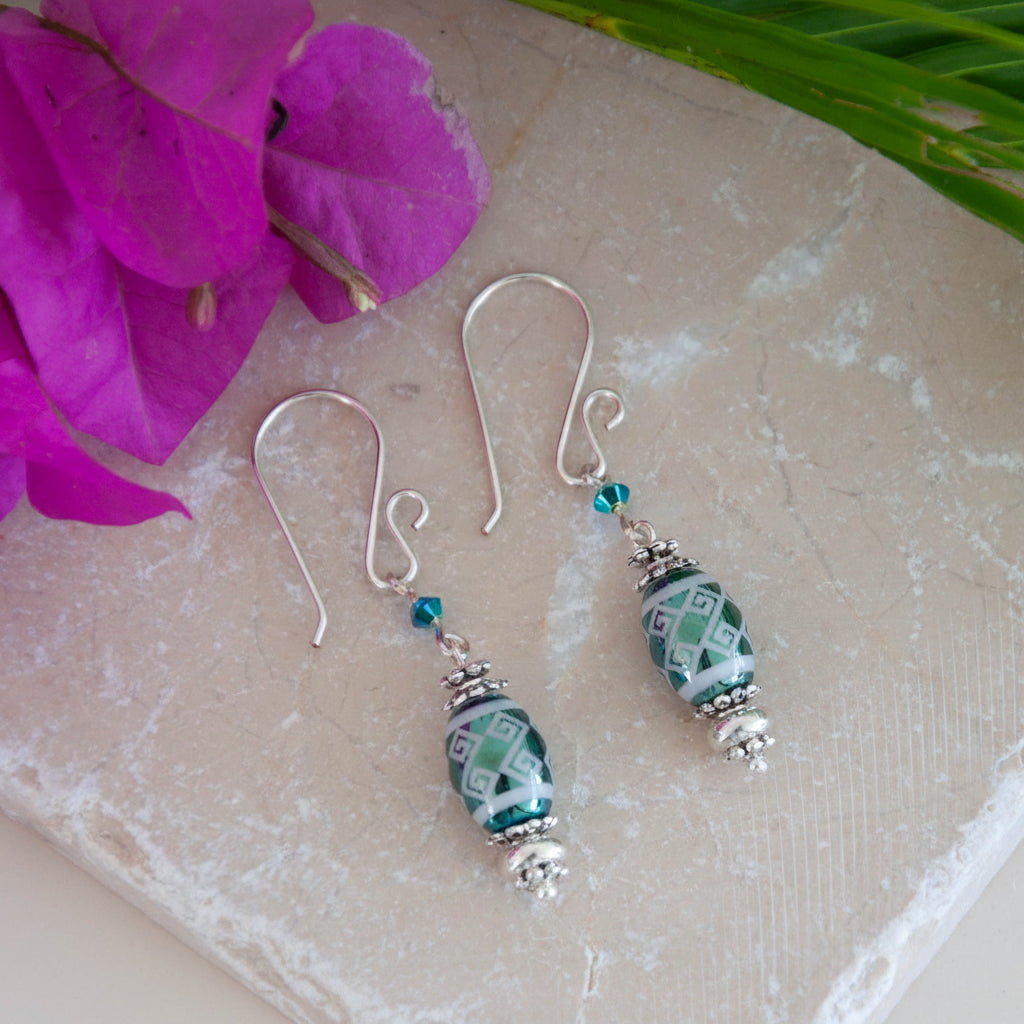Handmade teal metallic laser etched crystal rice bead drop earrings with tribal stripe pattern, Swarovski bicone crystal, silver-tone Bali spacer beads, and handcrafted silver-tone French hook ear wires, laid on travertine tile with a magenta flower and tropical palm leaves.