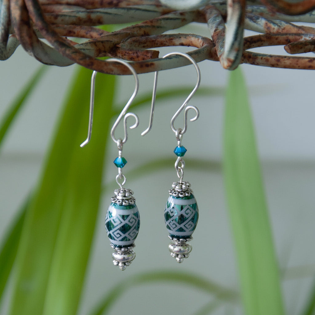 Handmade teal metallic laser etched crystal rice bead drop earrings with tribal stripe pattern, Swarovski bicone crystal, silver-tone Bali spacer beads, and handcrafted silver-tone French hook ear wires, hanging from a rustic wire branch with tropical green leaves.