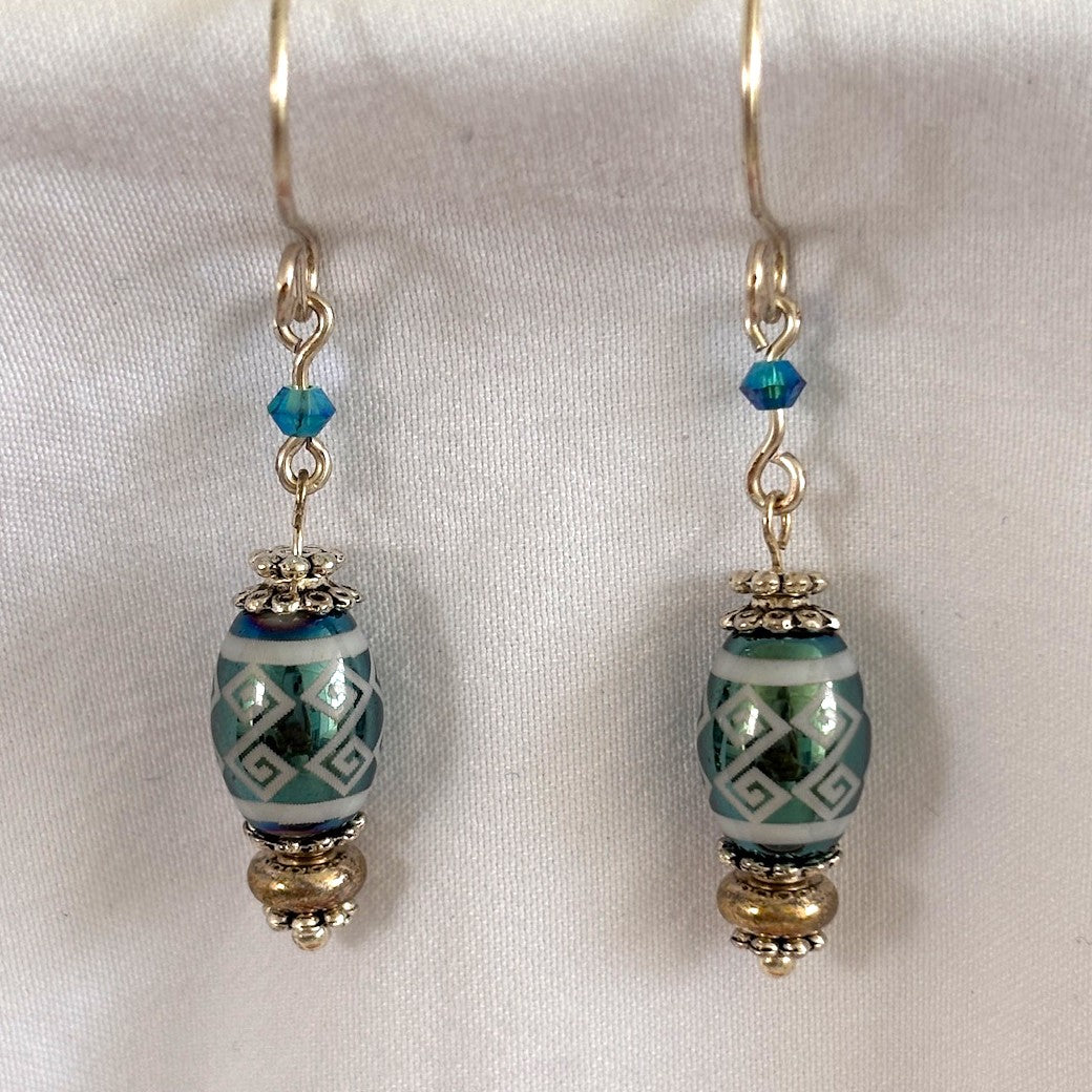 Pair of handmade teal metallic laser etched crystal rice bead drop earrings (8mm × 10mm) with geometric tribal stripe pattern, 3mm teal Swarovski bicone crystals, silver-tone Bali-style daisy spacer beads, and handcrafted silver-tone French hook ear wires, displayed on white fabric background.
