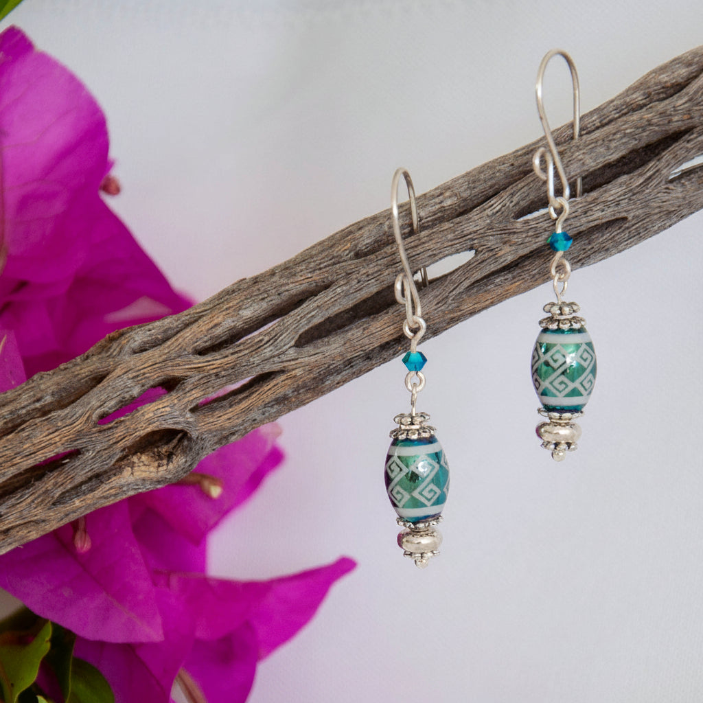 Handmade teal metallic laser etched crystal rice bead drop earrings with tribal stripe pattern, Swarovski bicone crystal, silver-tone Bali spacer beads, and handcrafted silver-tone French hook ear wires, hanging from driftwood with magenta bougainvillea.