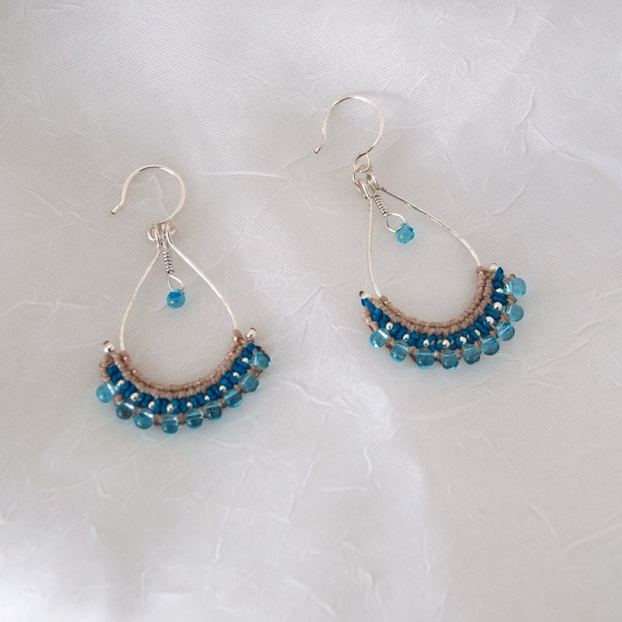 Pair of handmade hammered silver teardrop hoop earrings with light blue Miyuki teardrop fringe and teal macrame knotwork, displayed on white linen surface