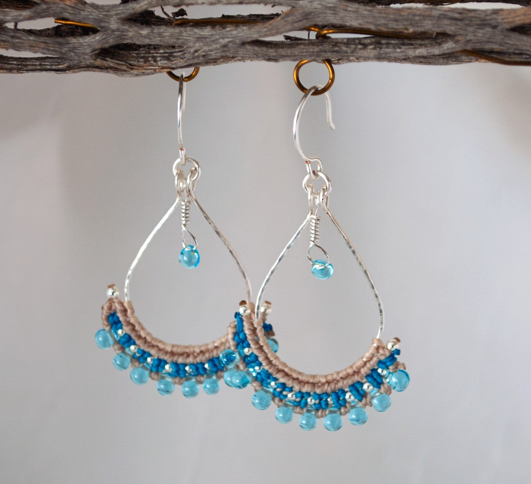 Handmade hammered silver teardrop hoop earrings with light blue Miyuki teardrop bead fringe, teal and beige macrame knotwork, and wire-wrapped teardrop charm, hanging from driftwood against a neutral background