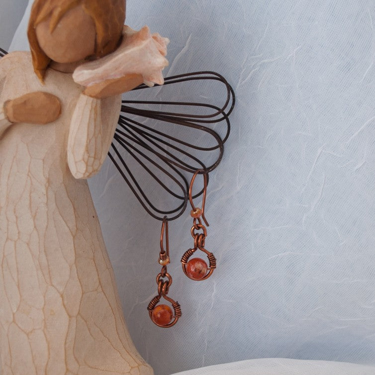 Handmade Ancient Cellar agate drop earrings with antiqued copper wire halo and iridescent orange crystal bead, displayed on Willow Tree angel figurine — artisan handcrafted boho earrings.