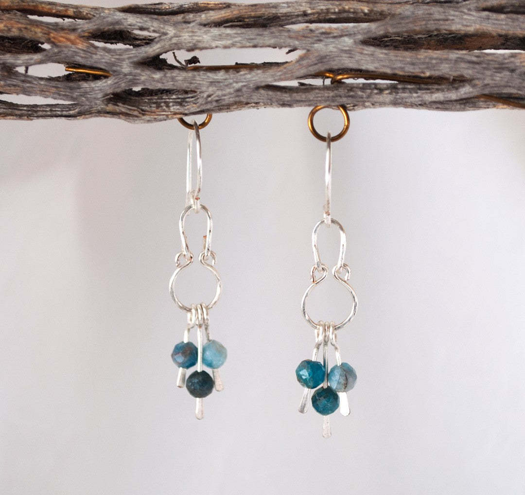 Apatite earrings with hammered silver oval and round frame chandelier structure and three faceted apatite bead dangles hanging from driftwood against gray-white background