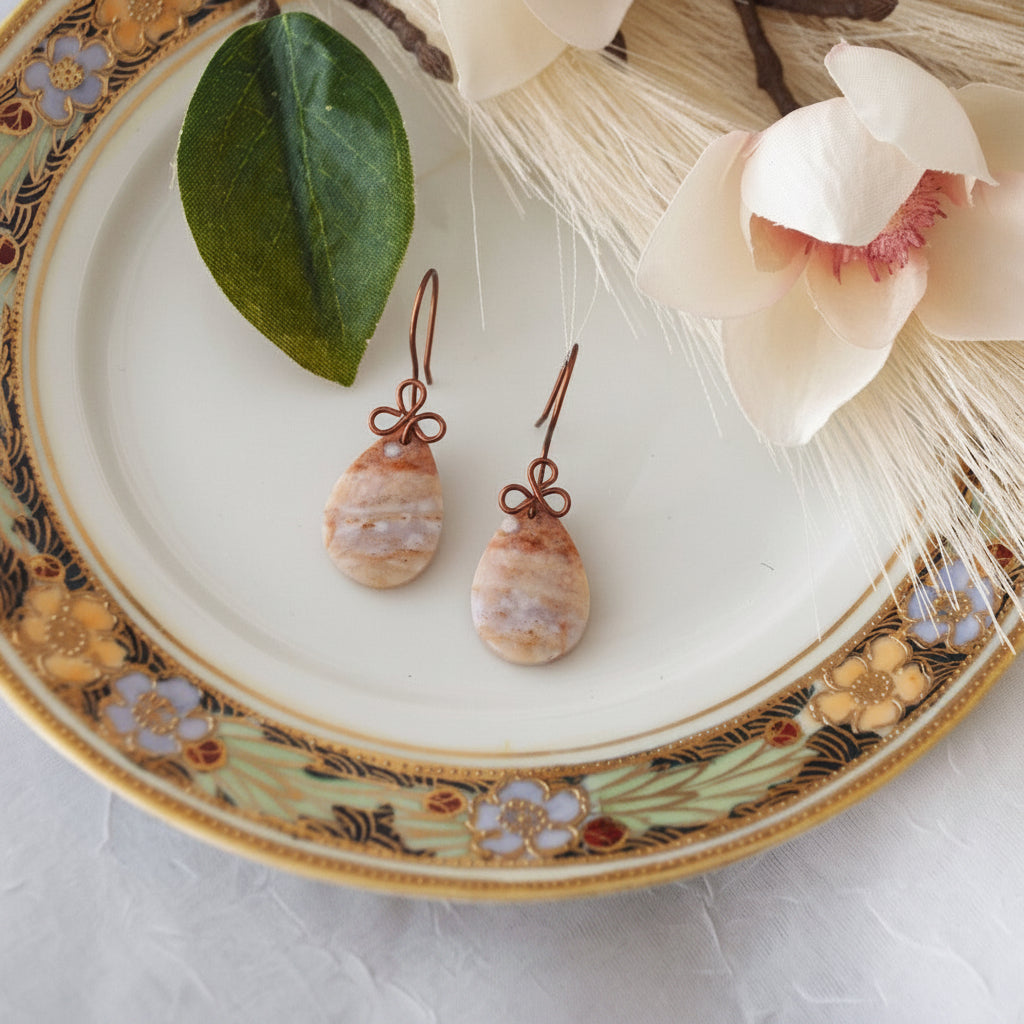 Handmade ocean jasper cabochon drop earrings with 15x26mm smooth teardrop ocean jasper and handcrafted antique copper wire bow, displayed on a vintage floral china plate with magnolia petal, green leaf, and pampas grass.