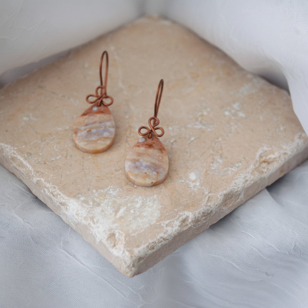 Handmade ocean jasper cabochon drop earrings with 15x26mm smooth teardrop ocean jasper and handcrafted antique copper wire bow, displayed on a travertine tile against a white linen background.
