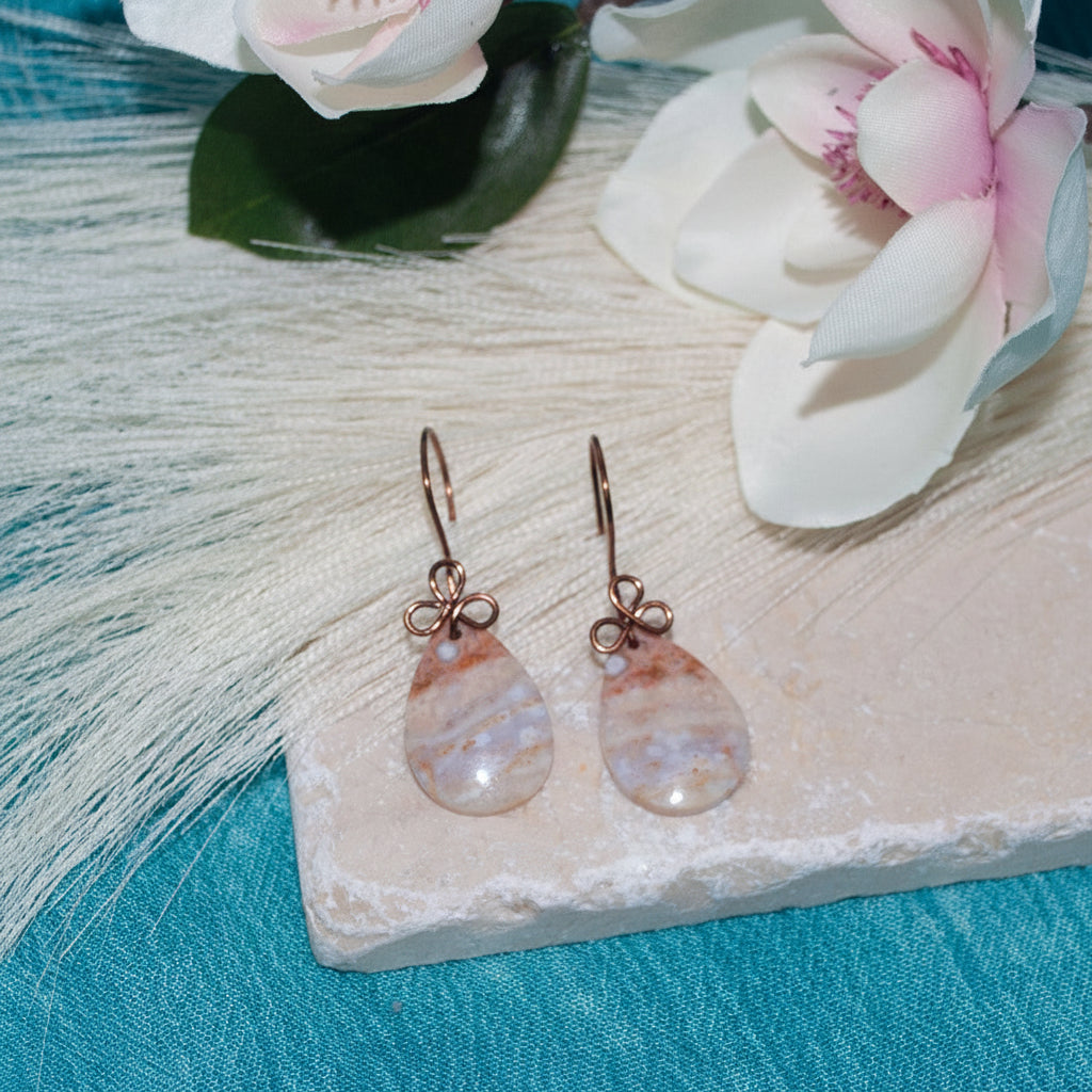 Handmade ocean jasper cabochon drop earrings with 15x26mm smooth teardrop ocean jasper and handcrafted antique copper wire bow, displayed on travertine tile with white and pink magnolia flowers and teal backdrop.
