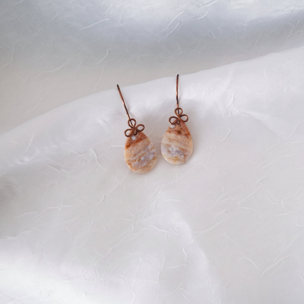 Close-up of handmade ocean jasper cabochon drop earrings with 15x26mm smooth teardrop ocean jasper and handcrafted antique copper wire bow, laid on white linen fabric.