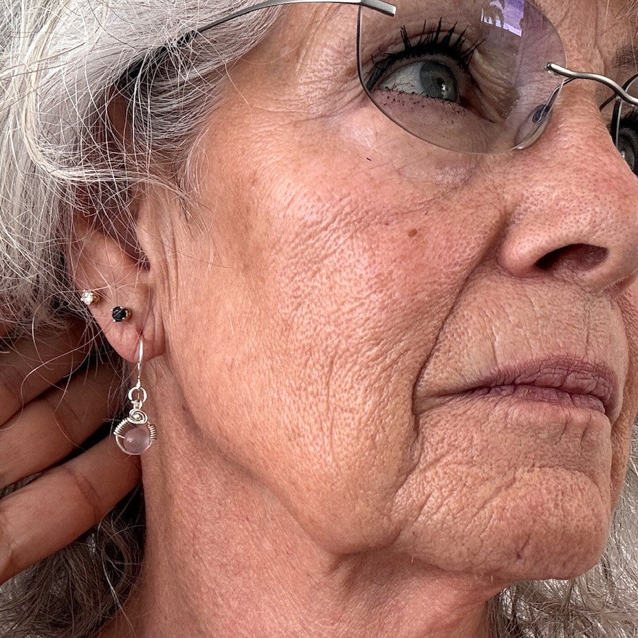 Pink cat's eye earring with silver-plated wire halo and wire-wrapped rose detail shown worn on model with silver hair and glasses