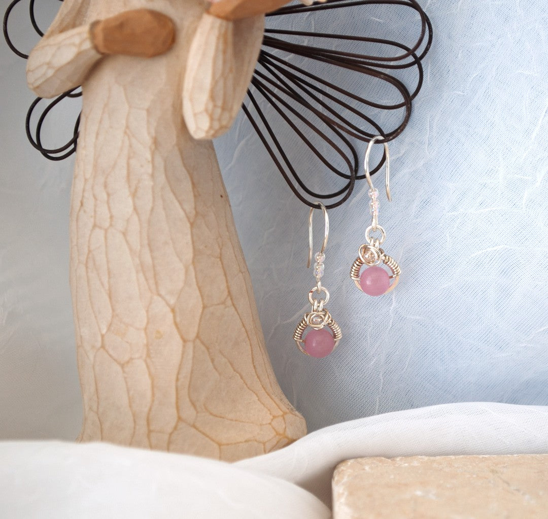 Pink cat's eye earrings with handcrafted silver wire halo and iridescent seed bead accents displayed on Willow Tree angel figurine against blue-gray background with travertine