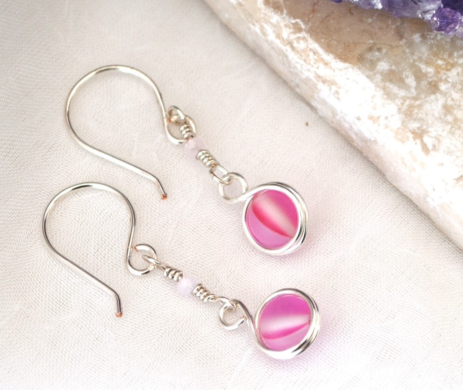 Handmade pink mermaid glass earrings with 6mm double wire-wrapped pink mermaid glass bead and faceted light pink crystal rondelle accent on silver-plated ear wires, displayed on white linen surface beside raw amethyst crystal cluster
