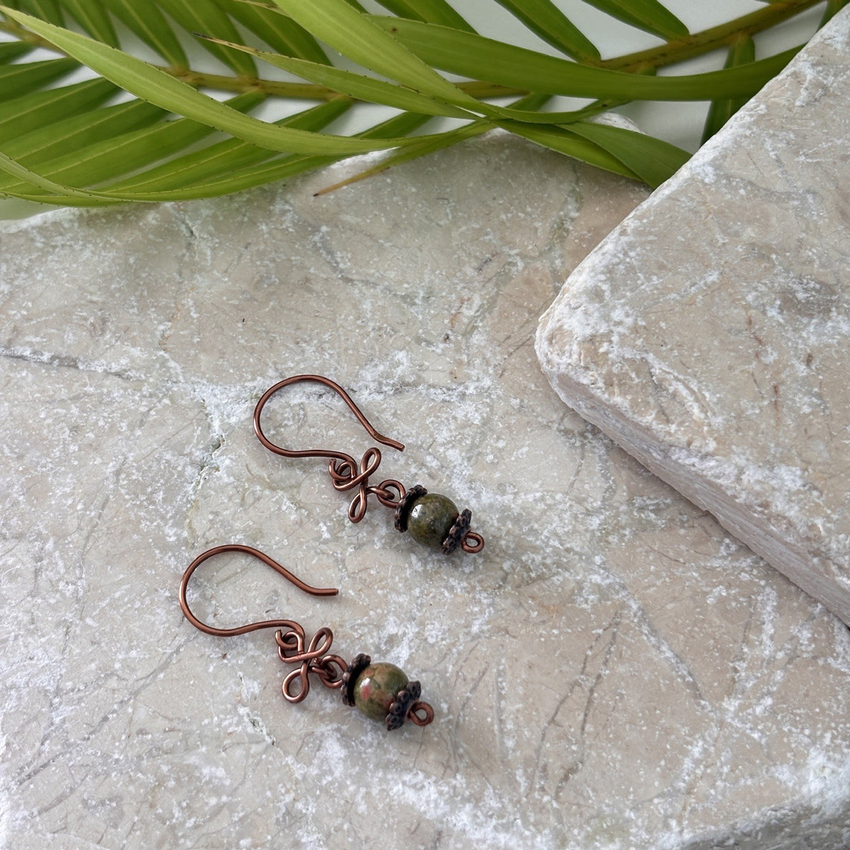 Pair of handmade unakite gemstone drop earrings with antique copper clover charm, displayed flat on travertine stone with tropical palm leaves
