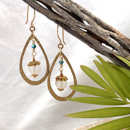 Mermaid glass earrings with hammered brass teardrop frame and blue-green Swarovski bicone hanging from driftwood with palm leaf and white linen background