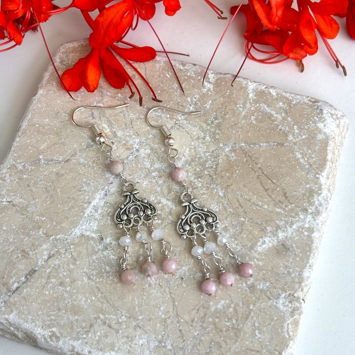 Handmade pink opal chandelier earrings with ornate silver-tone filigree and three beaded paddles displayed on travertine surface with red tropical flowers