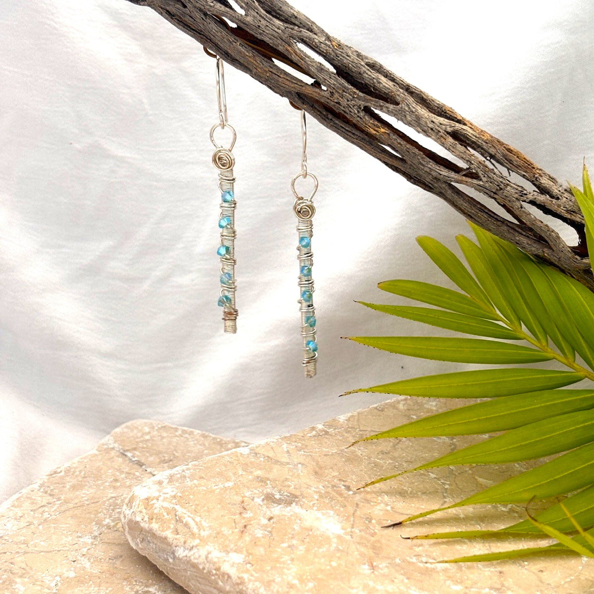 Aqua Swarovski earrings with wire-wrapped silver-plate bar and hand-coiled ring hanging from driftwood against white linen background with palm frond and travertine