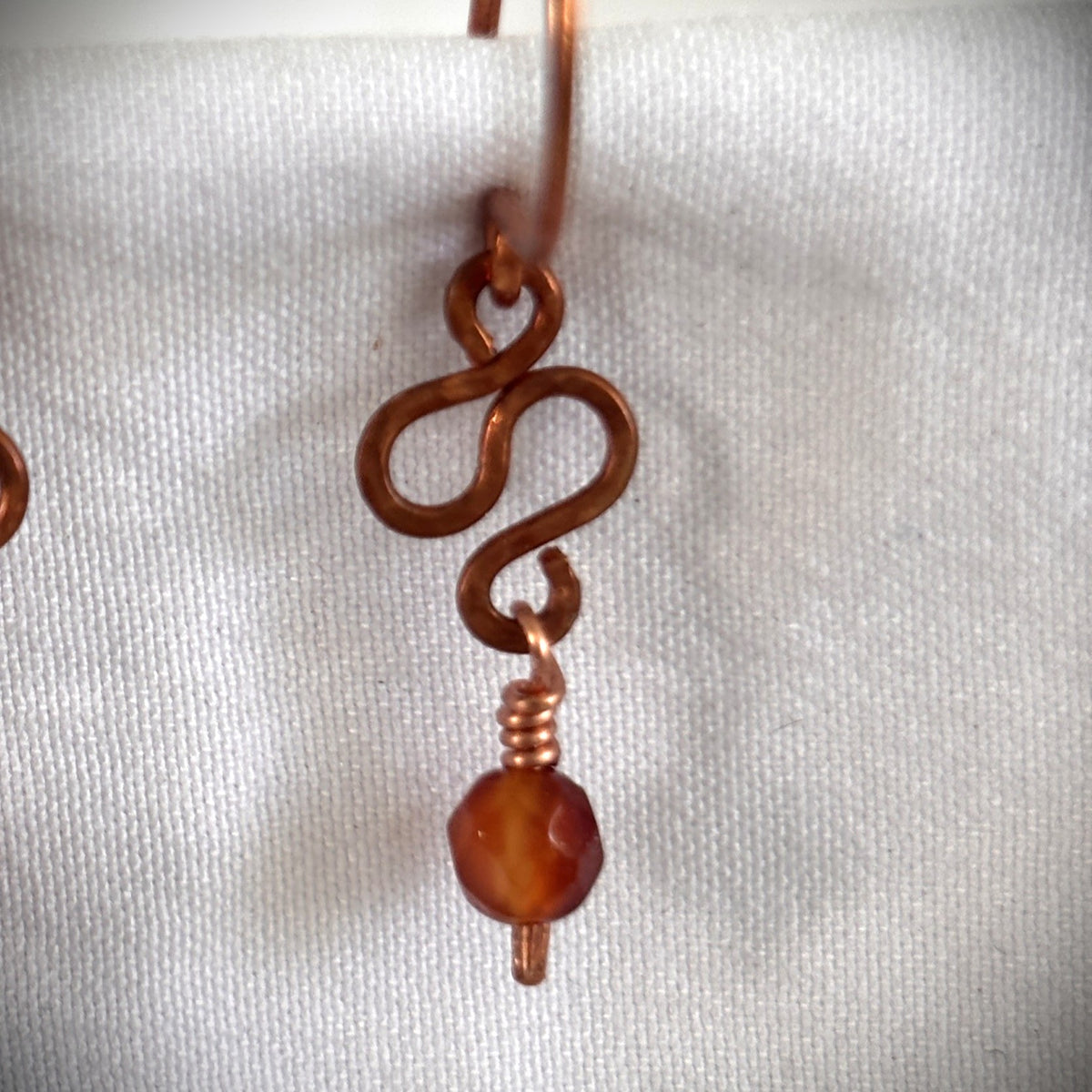 Close-up of handmade carnelian earring showing hand-formed hammered antique copper S-curve detail and wire-wrapped 4mm faceted carnelian bead drop on white linen