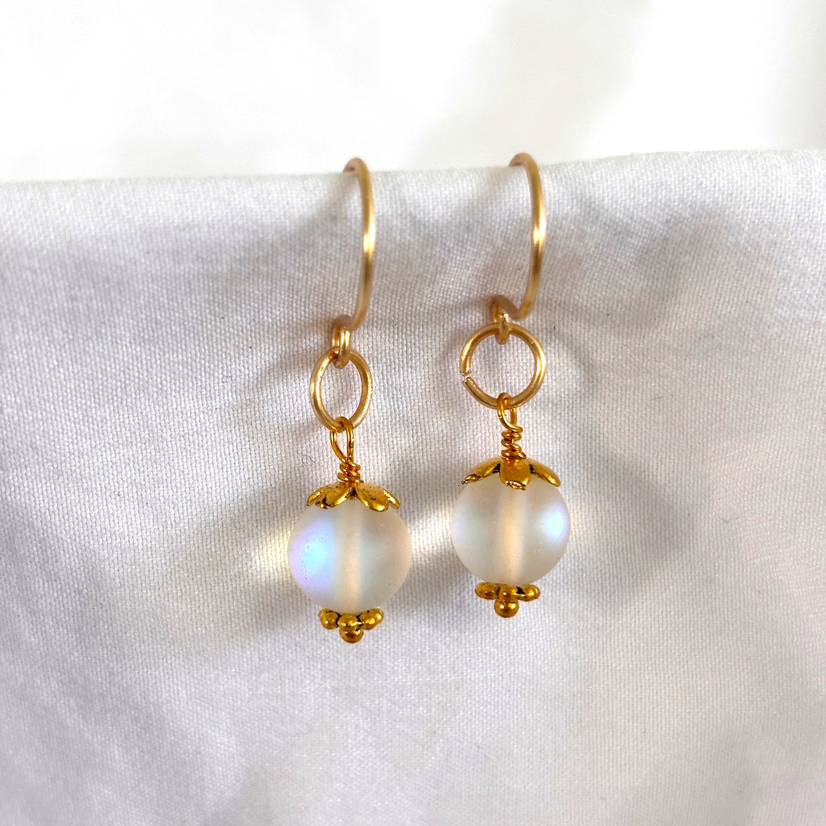 Handmade mermaid glass earrings with frosted iridescent 8mm bead, gold-tone bead cap and daisy spacer, oversized brass jump ring, and handcrafted brass French hook ear wire, displayed on white linen