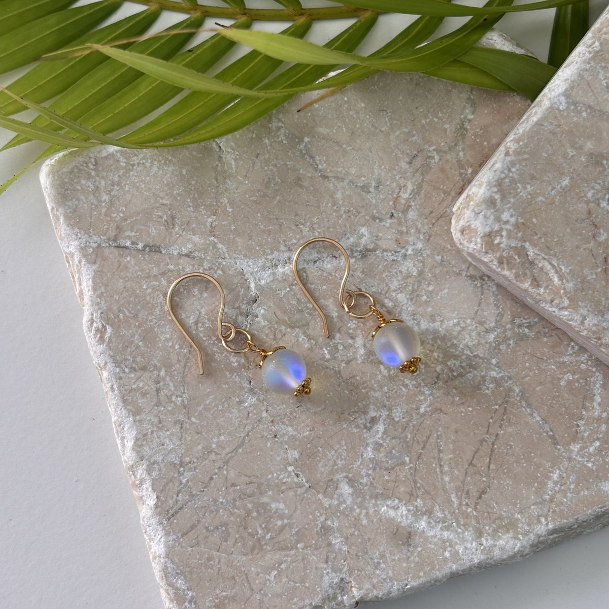 Handmade mermaid glass earrings with frosted iridescent bead, gold-tone bead cap and daisy spacer, and brass jump ring displayed on travertine surface with palm frond