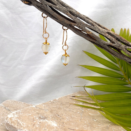 Frosted iridescent mermaid glass earrings with gold-tone bead cap and daisy spacer and oversized brass jump ring hanging from driftwood against white linen background with palm frond and travertine