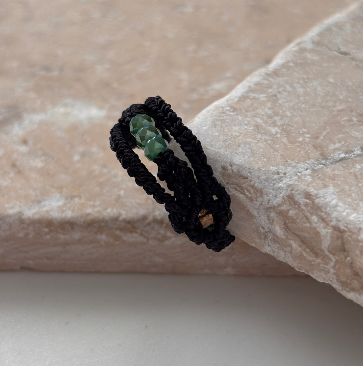 A side view of a handmade macrame band ring in black flat waxed nylon cord with a double-wrap braided design and three faceted mint green glass crystal beads with a square gold-tone seed bead accent visible on the underside, leaning against a pink stone surface against a white background,
