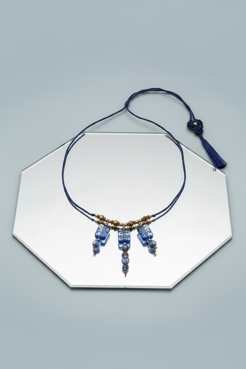 Handmade cobalt blue lampwork glass macrame necklace with gold foil detail displayed on hexagonal mirror showing adjustable cord, hematite bead accent, and handcrafted blue tassel closure.