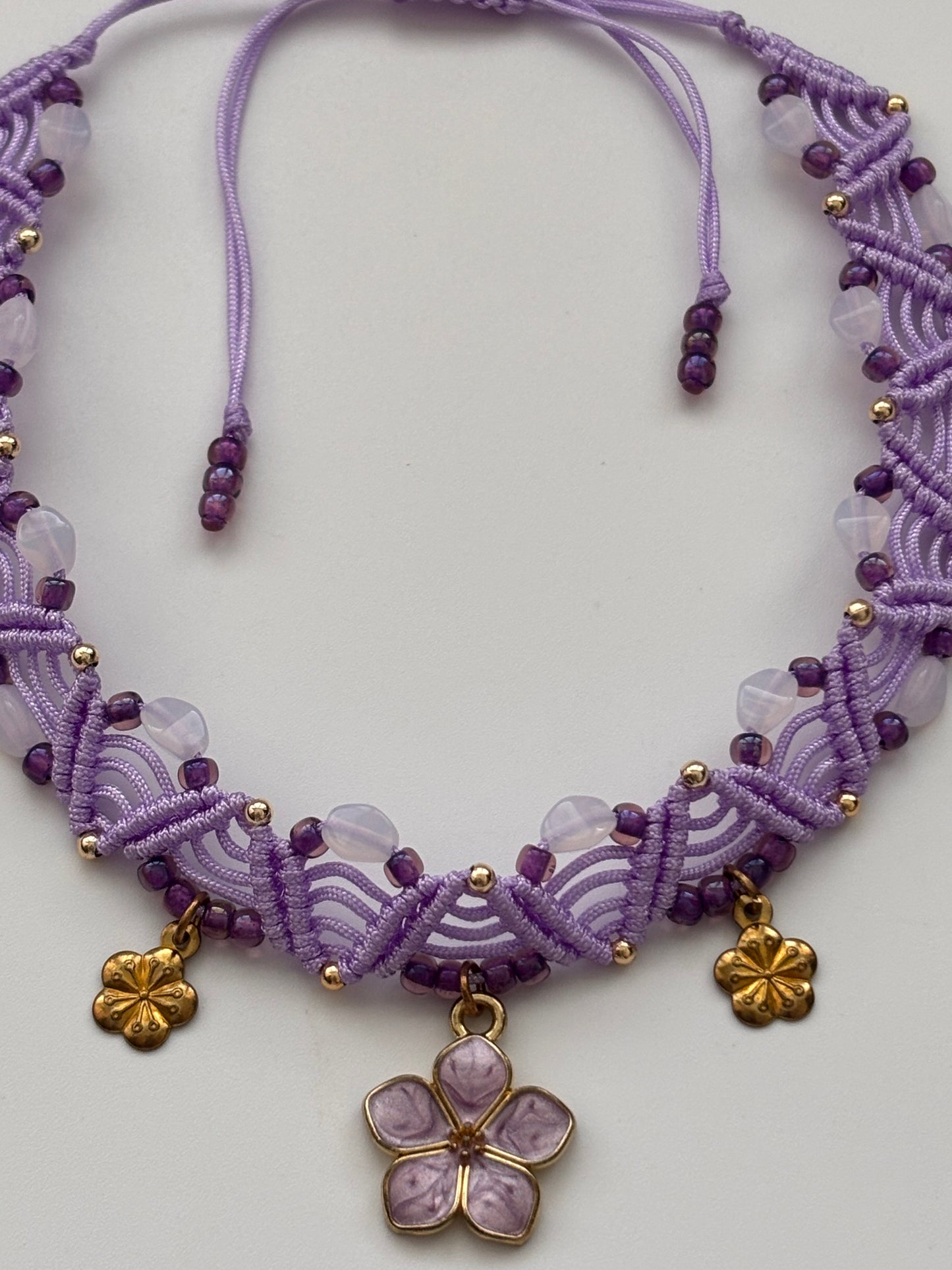 A purple macrame-style beaded anklet with a central lavender flower charm and two small gold flower charms, displayed on a light gray background.