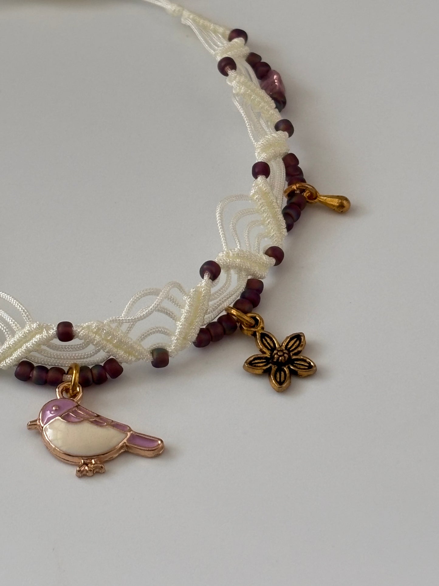 A close-up of a handmade ivory macrame anklet with dark plum matte seed beads, a lavender and white enamel bird charm, a gold-tone flower charm, and a gold-tone water drop charm, displayed on a neutral light gray background.