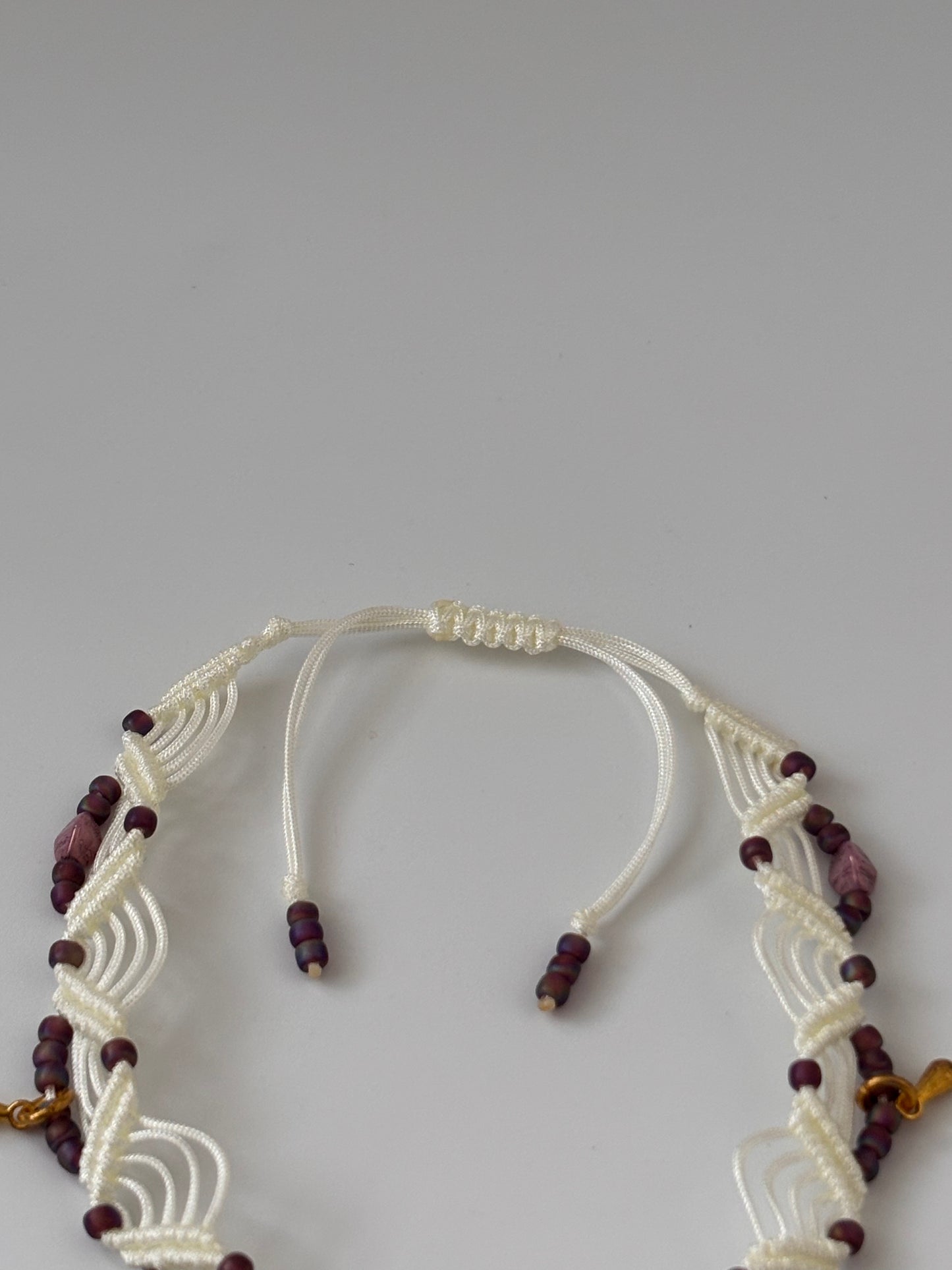 A close-up of the adjustable slide closure on a handmade ivory macrame anklet, showing the double zig-zag knotwork and dark plum matte seed beads on a light gray background
