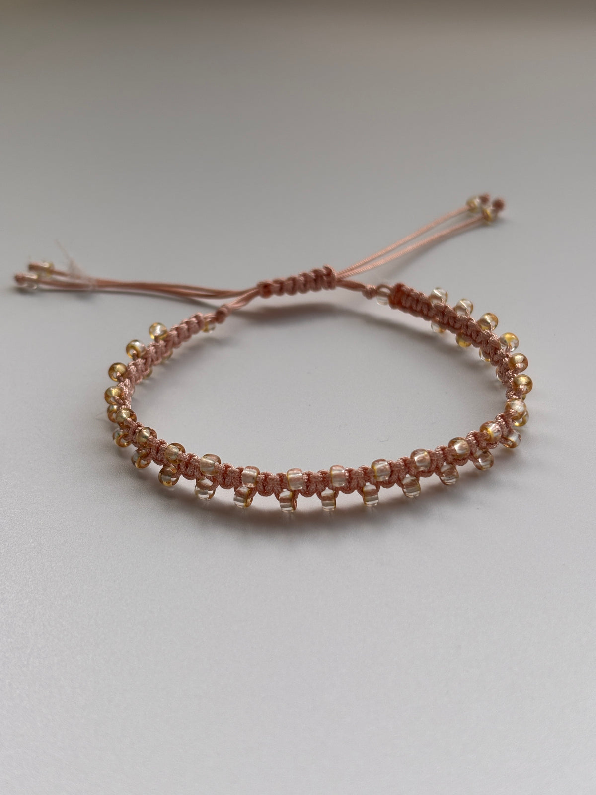 A handmade beaded macrame bracelet with copper edged seed beads, featuring an adjustable cord closure, displayed against a plain light gray background.