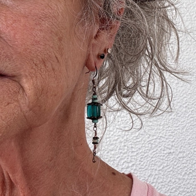 Handmade teal dyed glass drop earring with copper filigree caps, sea sediment beads, and wire-wrapped copper waterdrop charm shown worn on model with silver hair.