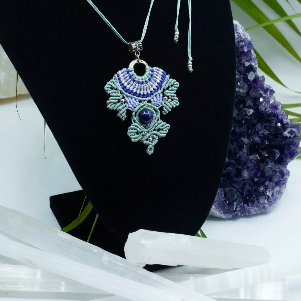 A handmade macrame pendant necklace with a blue and green woven design, displayed on a black jewelry bust. The necklace features a round Amethyst bead and is styled with purple amethyst crystals and white crystal points on a white surface.
