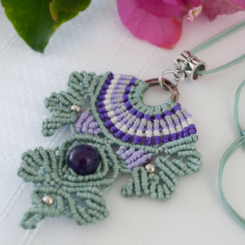 A handmade macrame pendant necklace with a green, purple, and white pattern, featuring an round Amethyst stone in the center and silver-tone beads, shown on a white textured background with pink flowers.