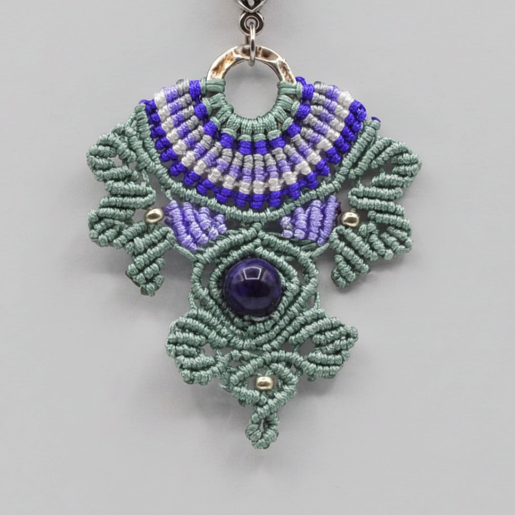 A handmade macrame pendant necklace featuring a green macrame-style leaf design with light and dark purple and white macrame accents, a central Amethyst stone, and a silver-tone ring clasp, shown on a neutral gray background.