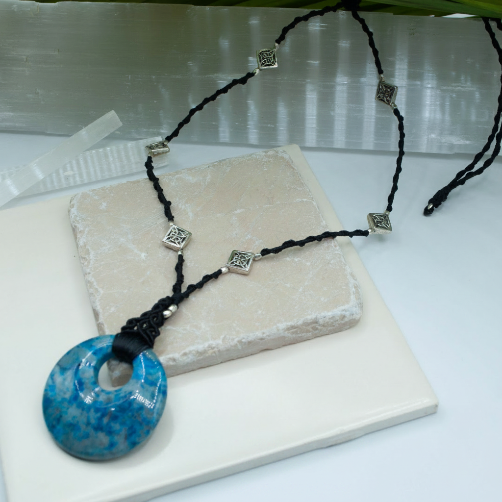 A black macrame cord necklace with silver-toned decorative beads and a large blue crazy lace agate donut pendant, displayed on a light stone tile over a white surface.
