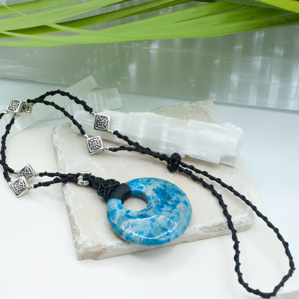 A blue crazy lace agate stone pendant necklace with a black macrame cord and silver-toned spacer beads, displayed on a white surface with a white selenite crystal and green leaves in the background.