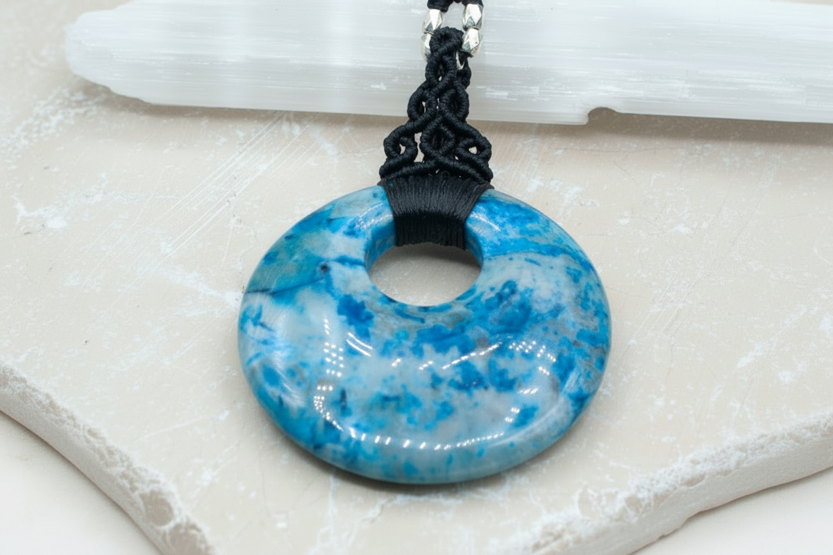 A blue crazy lace agate stone donut pendant necklace with a black macrame bail cord and silver metal spacer beads, shown close-up on a light stone surface.